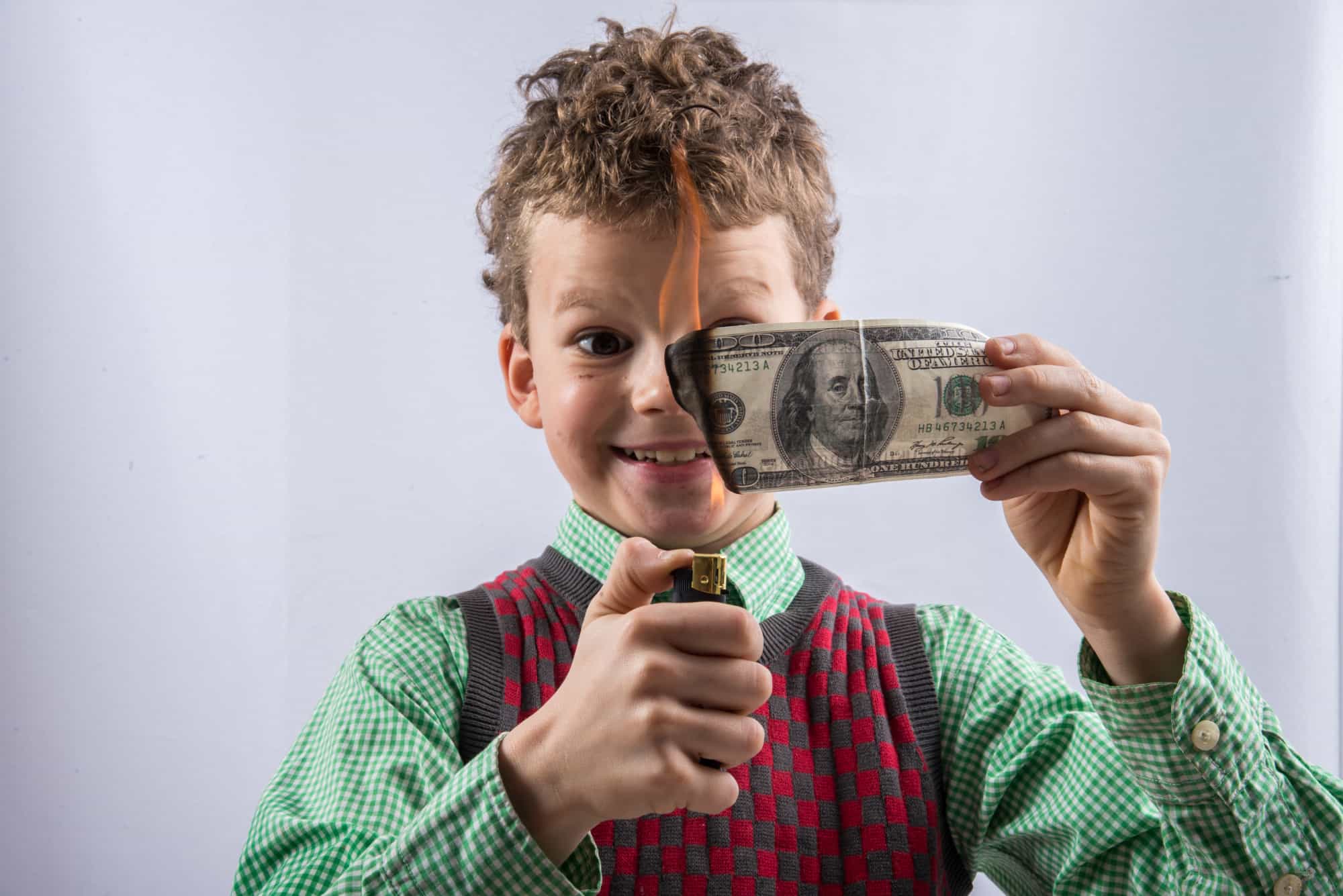 Redhead boy wearing green button-down shirt under red and gray sweater-vest burning a $100 bill with a lighter